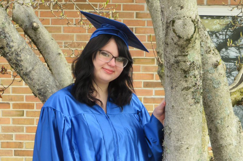 Wearing a blue graduation cap and gown, Skylyn smiles at the camera. 