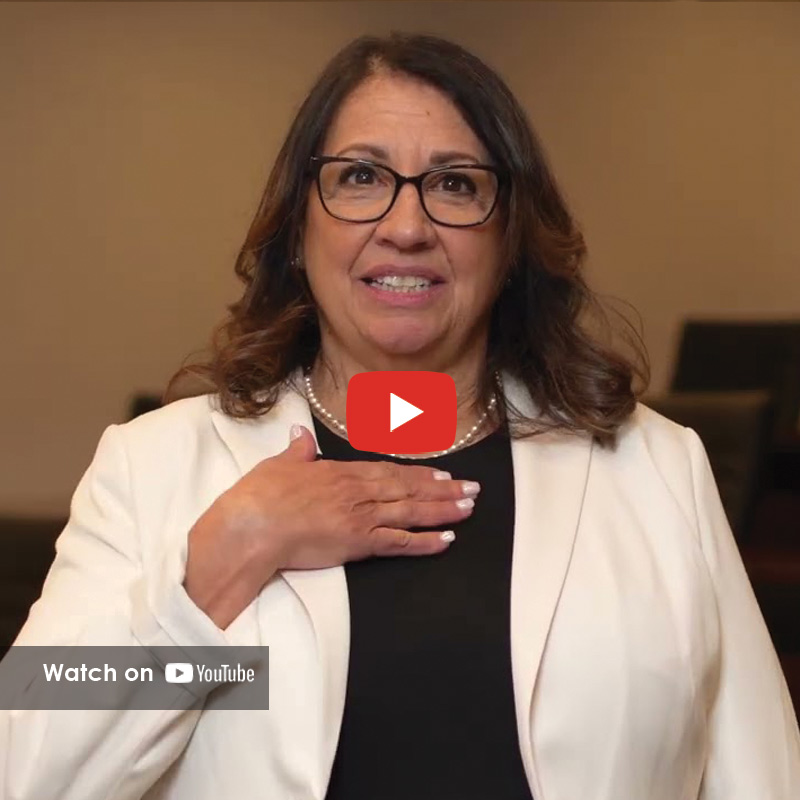 Woman with glasses speaking indoors, hand on chest, sharing a personal story