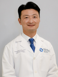 Man with short wavy black hair wearing a blue tie in a white lab coat