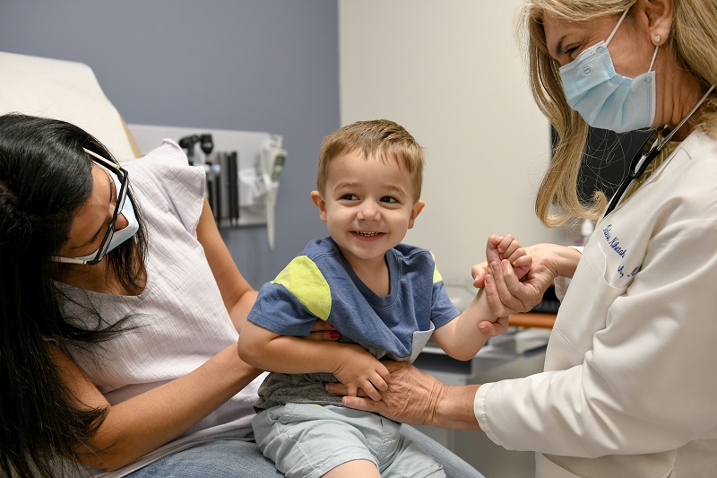 little boy smiling, sitting on mother's lap while interacting with the pediatrician