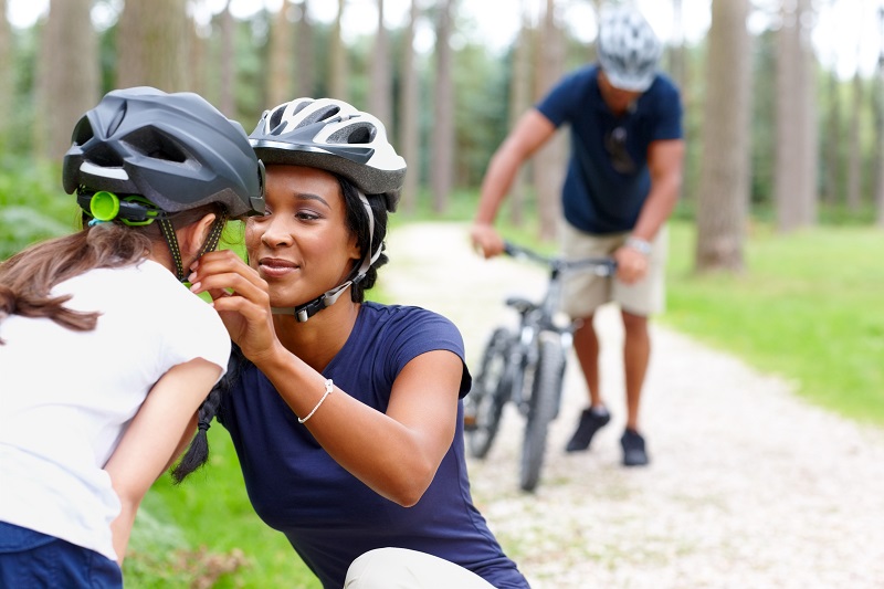 Caring young mother helping daughter put on helmet with father in background, wearing a helmet - Outdoors in country side