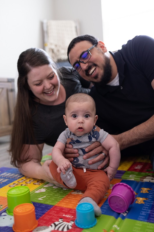Baby Benny sitting on a playmat with his toys, and his parents holding him, smiling.