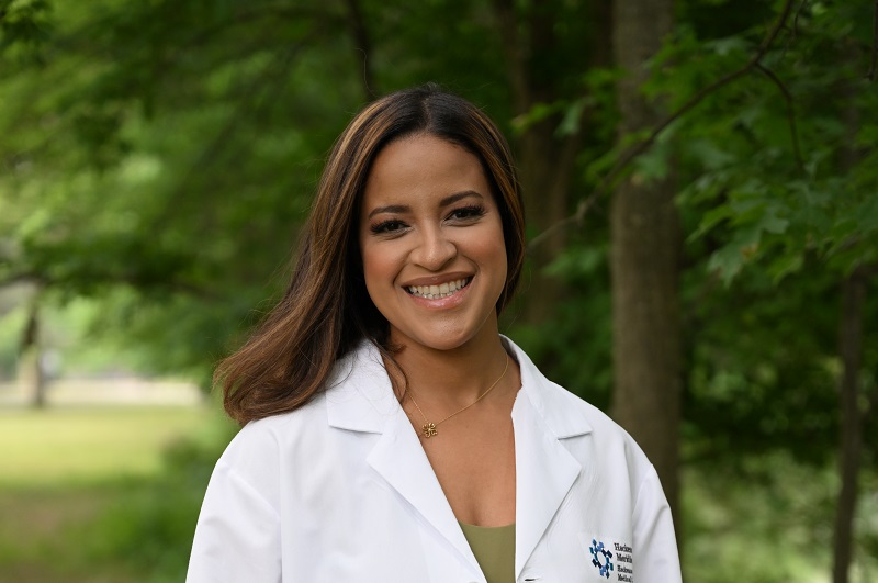 Dr. Magna Pastrano standing out in the park with, with her lab coat on, smiling, in front of trees.