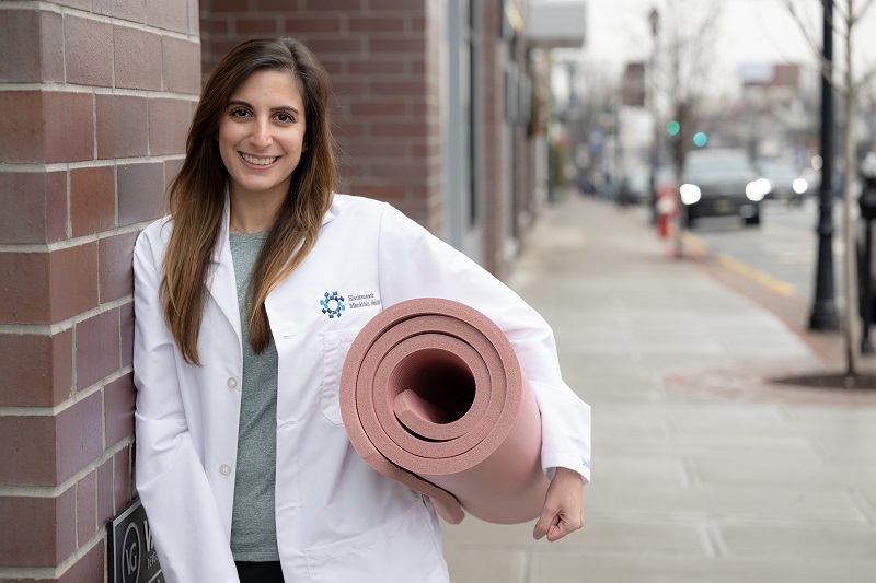 Dr. Sarah Mateen smiling and holding a rolled-up yoga mat.