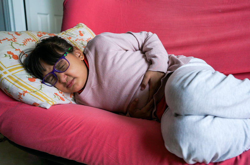Young girl with glasses on, clutching her stomach in pain from the stomach bug, laying on a pink couch.