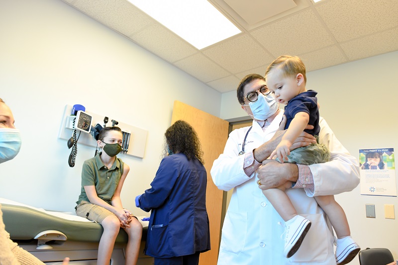 Pediatrician holding a child patient with their older sibling also sitting in the patient room. 