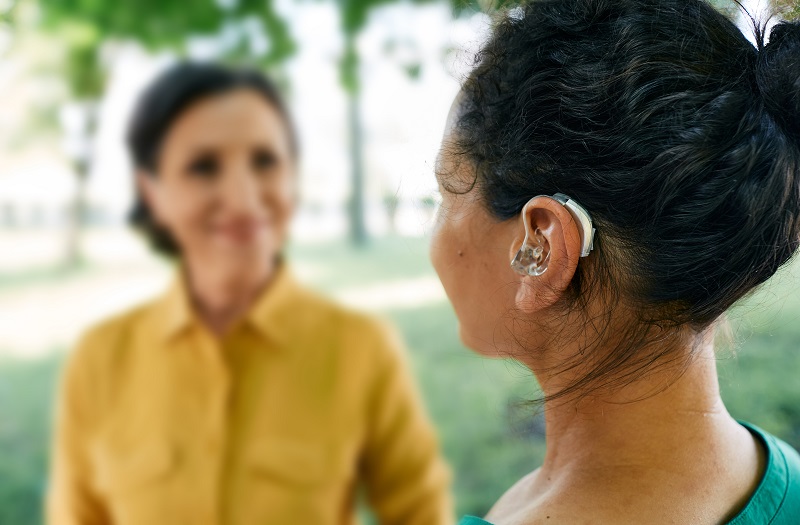 Adult woman with a hearing impairment uses a hearing aid to communicate with her female friend at city park.