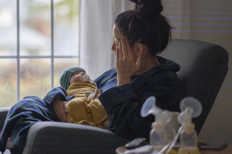 A new, postpartum mother sitting on a rocking chair in her living room wearing a housecoat holding her newborn baby. There is a breast pump and nursing supplies around.