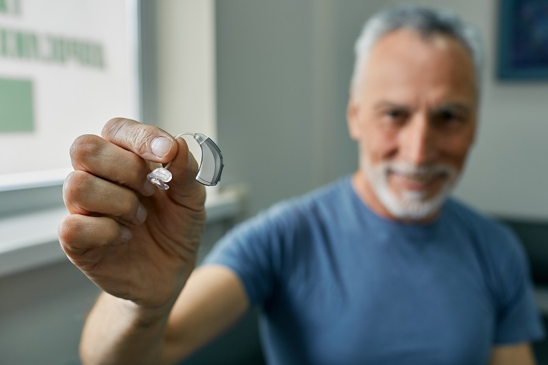 Senior man holding over-the-counter hearing aid in hand on foreground, close-up. Treatment of deafness in elderly people