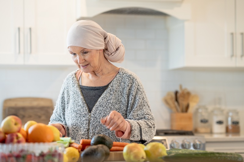 A senior woman with cancer stands at her kitchen counter as she works to prepare an assortment of fruits and vegetables. She is dressed casually in a sweater and wearing a headscarf as she looks over the produce and makes an effort to maintain a healthy diet during her Chemotherapy