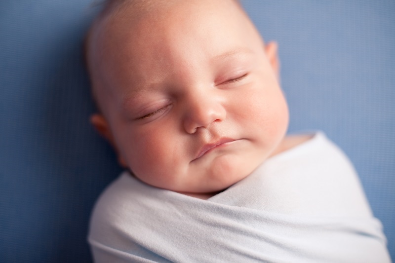 Close up of a sleeping baby lying on his back, swaddled, on a blue sheet.