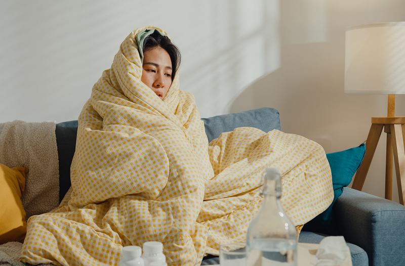 Young woman sick on the couch, wrapped up in a blanket, shivering from the flu and body aches.