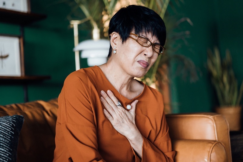 Woman with eyes closed holding her chest in discomfort, suffering from chest pain while sitting on the sofa at home.