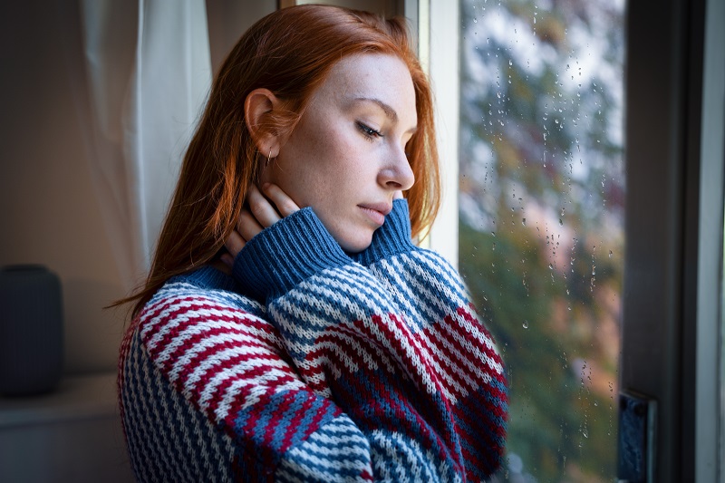 Woman looking out the window depressed as it rains and is darker outside, experiencing seasonal affective disorder.