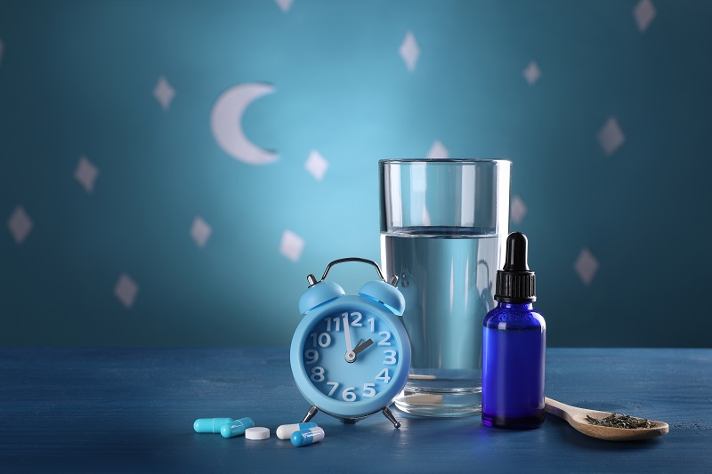 Alarm clock and different remedies for insomnia treatment near glass of water on table against blue wall decorated with stars and crescent