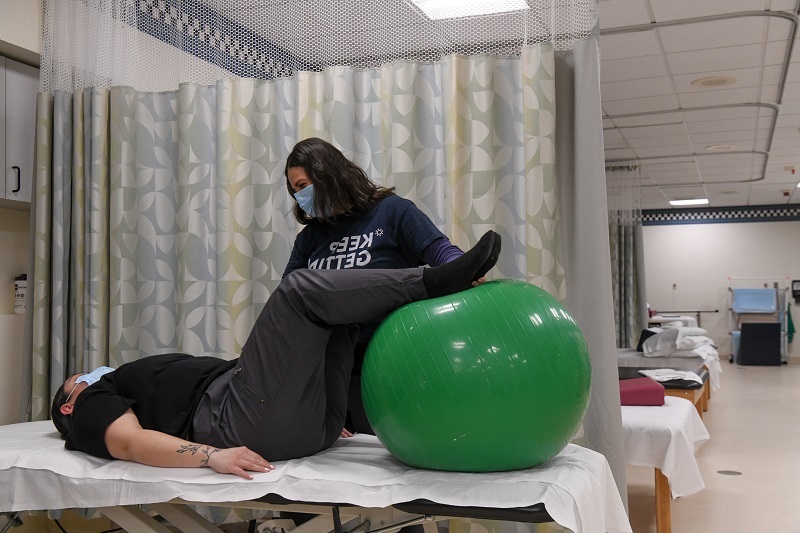 Physical therapist helps a patient do exercises with a large workout ball after a joint replacement. Patient is laying on the table with legs tabletop over the ball. 