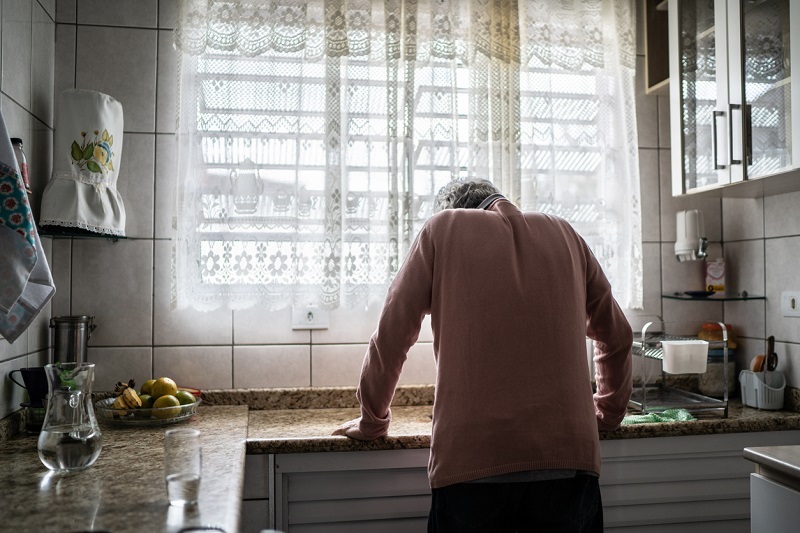 From behind, an older man leaning on a counter with fatigue, not feeling well.