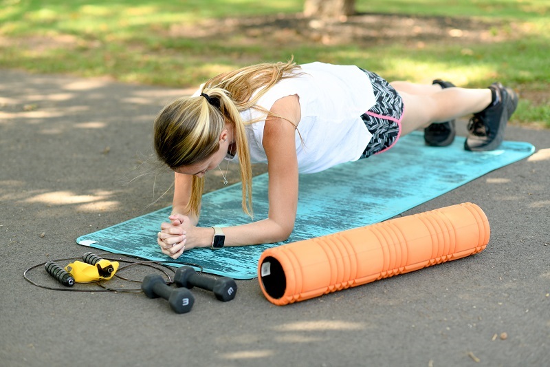 Woman doing a plank on a yoga mat outside in the park, with weights and a jump rope nearby. 