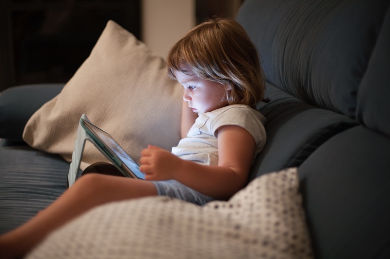 blonde three year old baby, sitting comfortably on the sofa inside home at night reading and watching digital tablet, face illuminated by the light of the screen