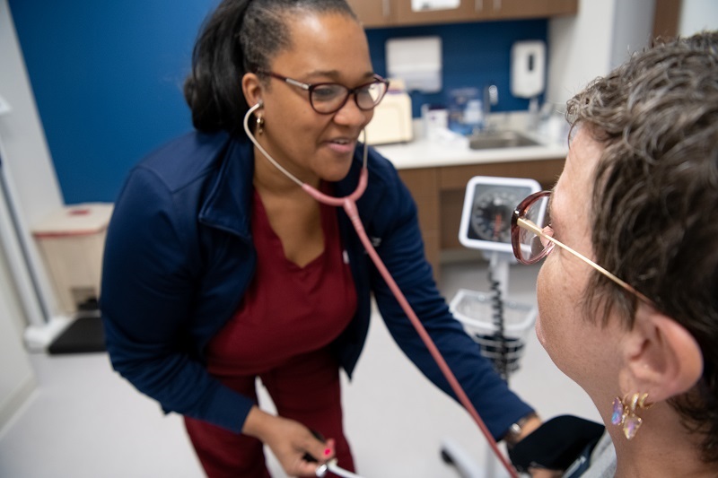 Woman at a primary care appointment, with a nurse leaning in to listen to her heart with a stethoscope.