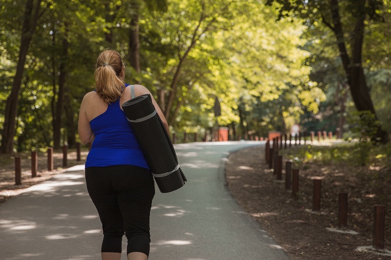 Woman walking away from the park and carrying exercising mat on her shoulders