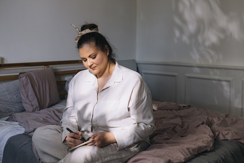 Woman sitting on her bed, in her pajamas taking note of her sleep.