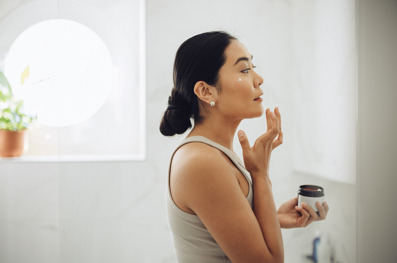 Woman standing in her bathroom, looking in the mirror, holding a jar of face cream and applying some to her face.