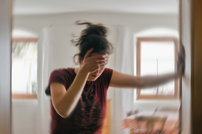 Woman standing, one hand on her forehead, one on the wall, trying to regain balance. Suffering
