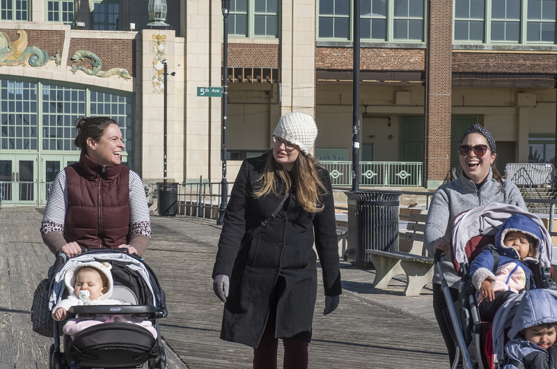 Three friends walking on the Asbury Park boardwalk, smiling, and pushing strollers with their children.