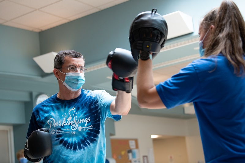 Patient with Parkinson's disease participating in Rock Steady Boxing program - working on coordination and strength by hitting hand targets. He is wearing a shirt that says ParkinSINGS.