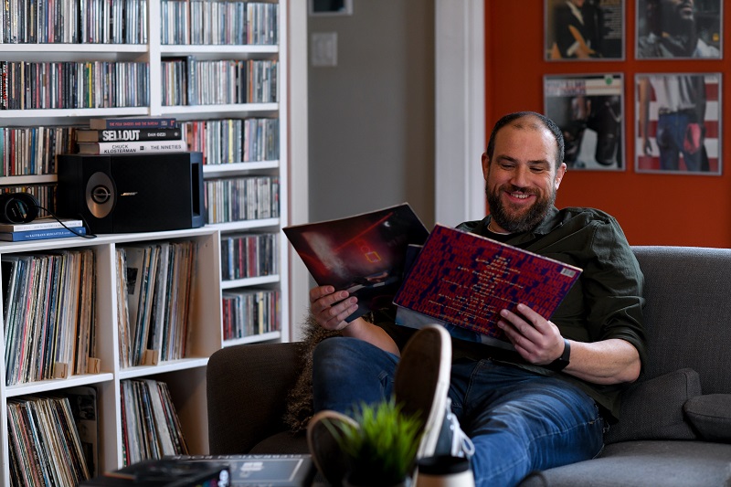 Matt Kleinschmidt, a young man, sitting on a couch holding vinyl records while smiling 