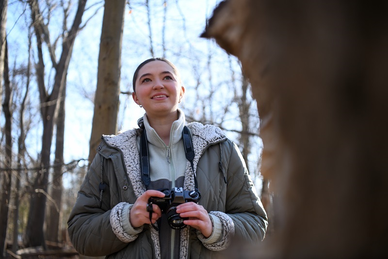 Young woman, Caitlyn, stands in the woods holding a camera, looking out and smiling.