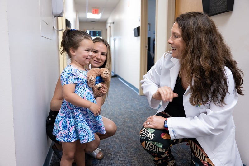 Toddler Maggie Hennessey smiling in the doctor's office hallway, with her mom and doctor.