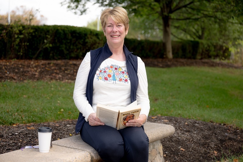 Debbie Cihoski sits on a bench in the park, reading a book and smiling.