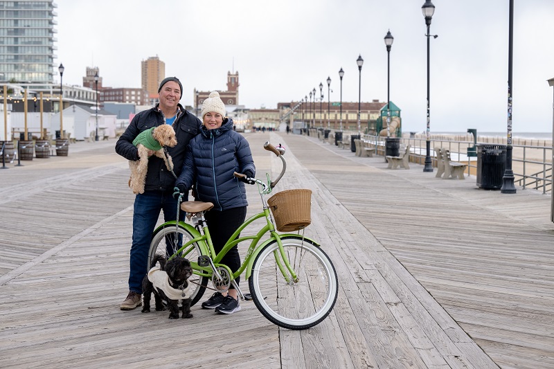 Cathy and her husband standing on the boardwalk in Asbury Park with her two dogs and bicycle.