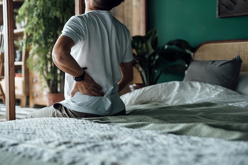 Rear view of a man sitting on the edge of the bed, holding his lower back in pain. 