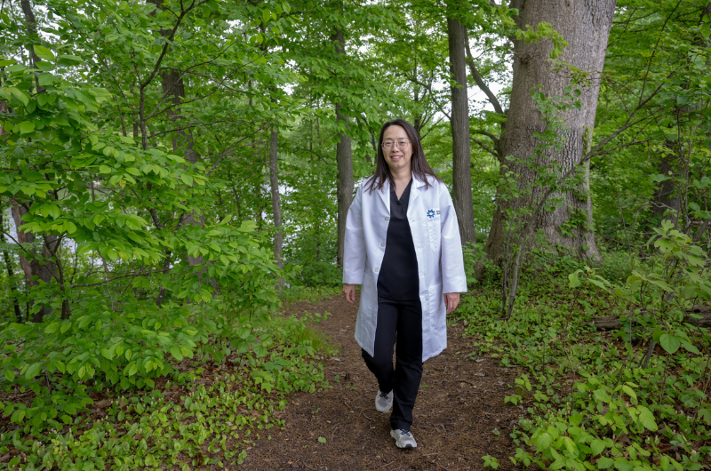 Xue Ao, M.D., stands in a wooded area surrounded by greenery, smiling at the camera.
