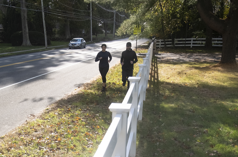 Woman and man running along the side of the road on soft grass.