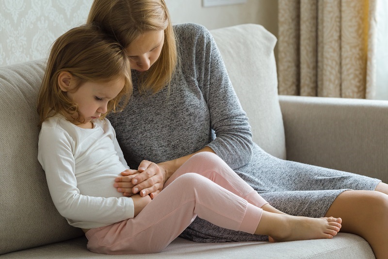 Child sitting on sofa and suffering from constipation, mother touching her belly trying to offer comfort.