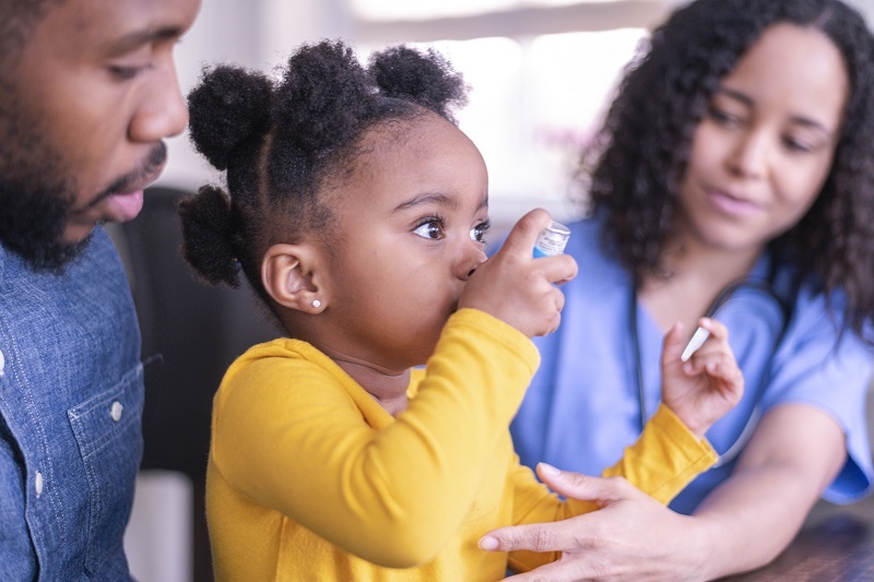 A little girl with asthma is at a doctor's appointment. She is sitting on her father's lap. The girl is using a puffer. The doctor is teaching the child how to use the medical device.