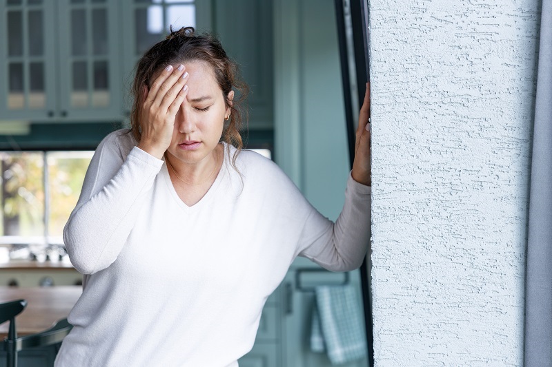 Woman with one hand on her forehead, the other resting on a wall for balance. Experiencing dizziness.