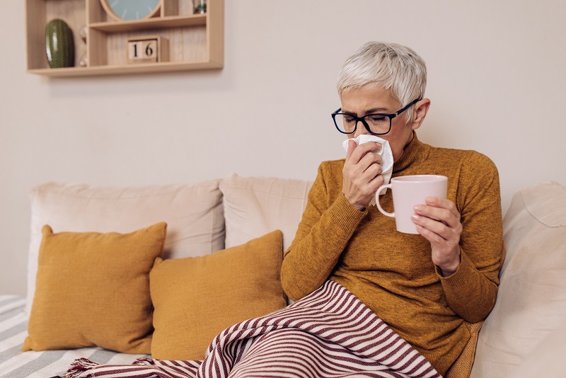 Older woman sitting on the couch, blowing her nose, holding a cup of tea.