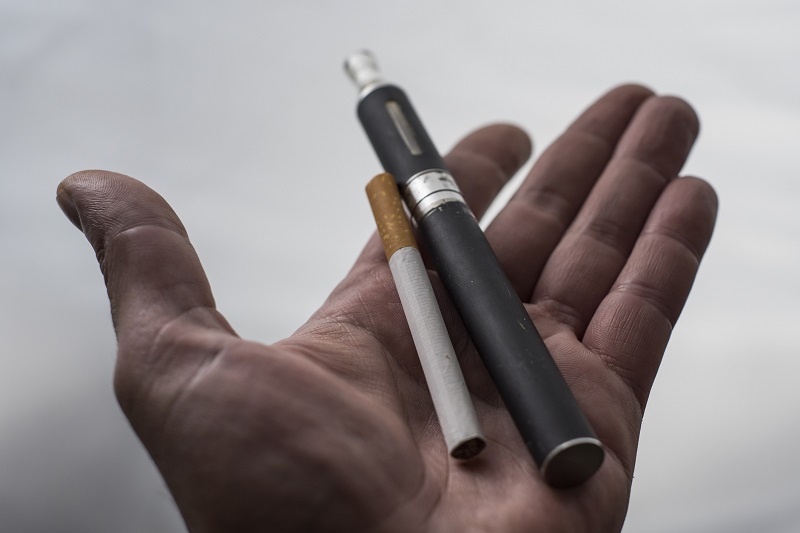 A man's hand holding a vaping cigarette and a traditional cigarette.