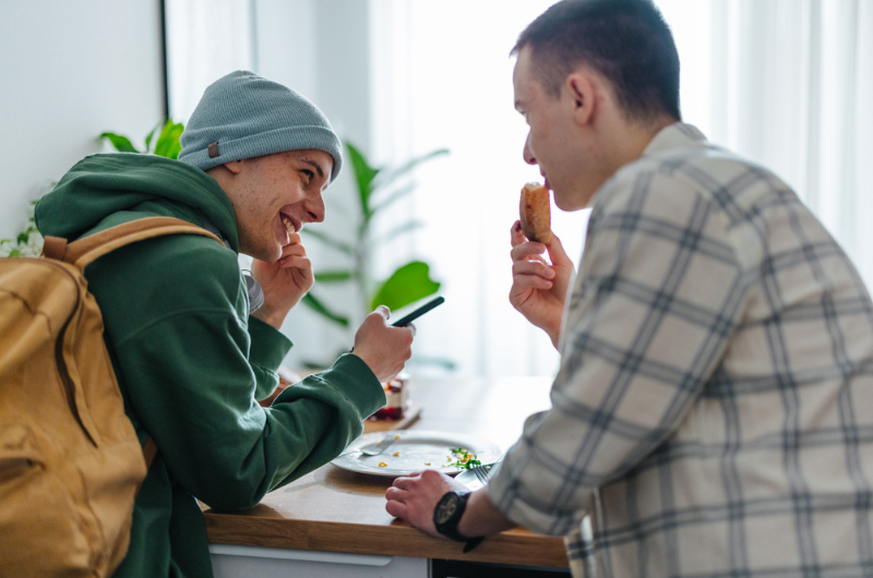 Two teenage boys eat food off a plate while smiling and laughing.