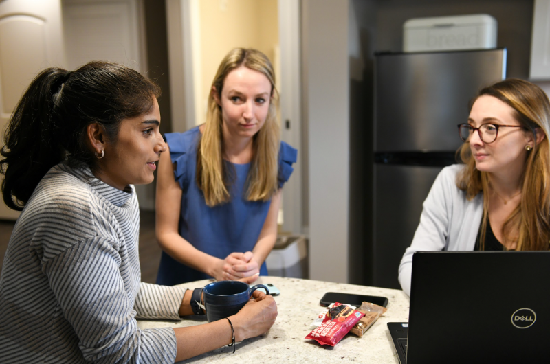 Three young women stand around a kitchen island, chatting.