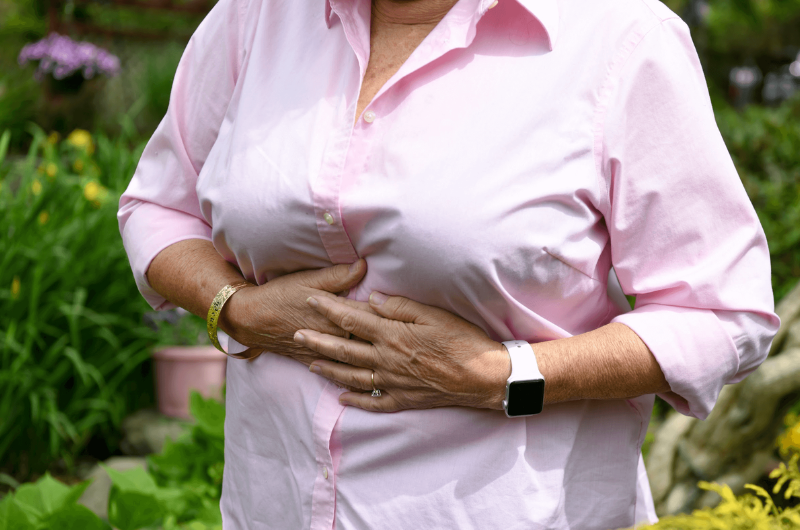 An older woman wearing a pink blouse clutches her upper stomach.