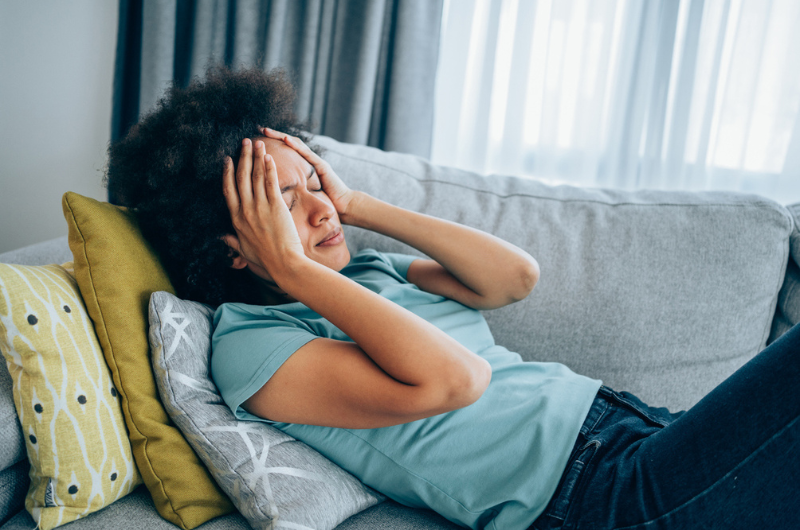 Young woman is lying on the couch and holding her head with hands.