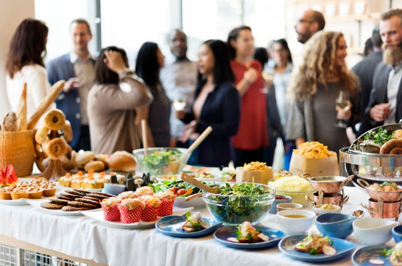 A buffet table full of food is shown with a small group of people in the background.