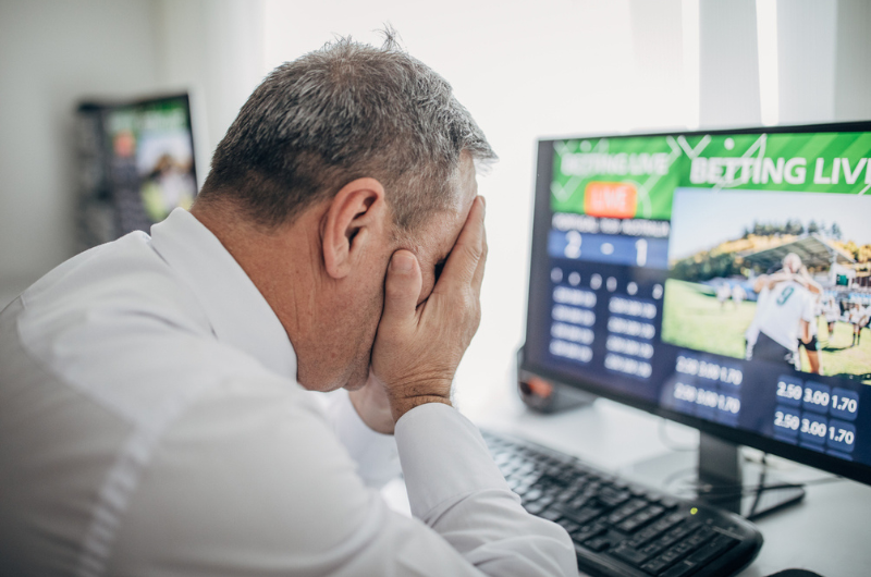 Older man with his face in his hands is doing sports betting using a computer.