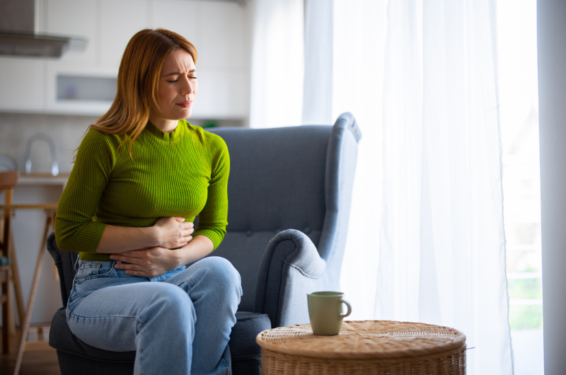 A young woman sits on a couch and clutches her abdomen in pain.